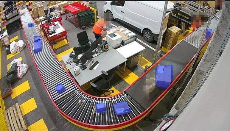 View of a conveyor in operation: totes on rollers, workstations, and operators' faces masked by the embedded processing.